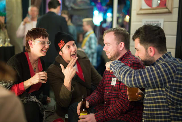 Group of four friends enjoying drinks and engaging in lively conversation at the Love Bomb 2017 event, with a festive atmosphere in the background.